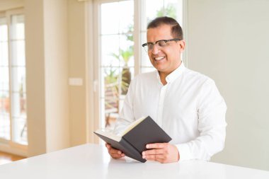 Orta yaş adam evde mutlu bir yüz ayakta gözlük giyen ve dişleri gösteren kendinden emin bir gülümseme ile gülümseyen bir kitap okuma