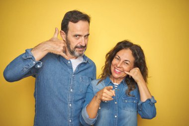 Güzel orta yaş çift birlikte izole sarı arka plan üzerinde duran telefon jest konuşma yapıyor gülümseyerek ve sizi işaret. Beni ara.