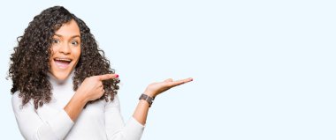 Kıvırcık saçlı Genç güzel kadın balıkçı yaka kazak hayran ve el ile sunmak ve parmak ile işaret ederken kameraya gülümseyen.