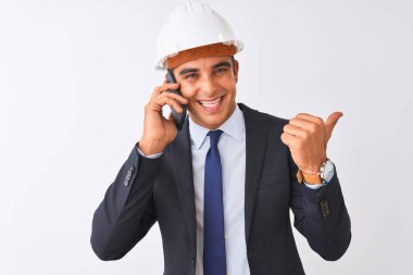 İzole beyaz arka plan üzerinde akıllı telefonda konuşan kask giyen genç mimar adam işaret ve mutlu yüzü gülümseyerek yan başparmak ile gösteren