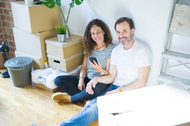 Orta yaş yaşlı çift ayakta mutlu bir yüz ile akıllı telefon kullanarak yeni bir eve hareket ve dişleri gösteren kendinden emin bir gülümseme ile gülümseyerek
