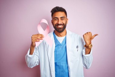 Genç hint doktor adam izole pembe arka plan üzerinde duran kanser kurdele tutan işaret ve mutlu yüzü gülümseyerek yan başparmak ile gösteren