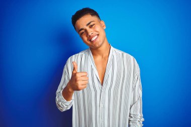 İzole mavi arka plan üzerinde duran çizgili gömlek giyen genç Brezilyalı adam el ile jest kadar mutlu başparmak yapıyor. Başarı göstererek kameraya bakan ifadeyi onaylama.