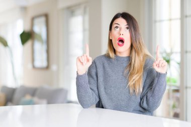 Evde kış kazak giyen genç güzel kadın hayrete ve yukarıya bakıyor ve parmakları ile işaret ve silah kaldırdı şaşırttı.