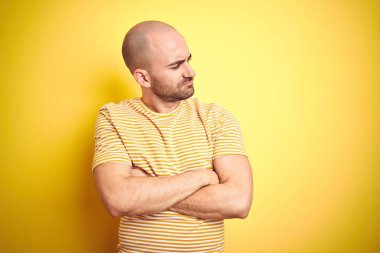 Sakallı genç kel adam sarı izole arka plan üzerinde rahat çizgili t-shirt giyen kolları ile yan bakarak ikna ve kendinden emin geçti