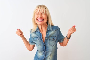 Orta yaş kadın izole beyaz arka plan üzerinde duran rahat kot gömlek giyen çok mutlu ve heyecanlı kolları kaldırdı ile kazanan jest yapıyor, gülümseyerek ve başarı için çığlık. Kutlama konsepti.