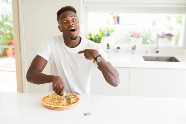African american man eating cheese pizza at home with surprise face pointing finger to himself