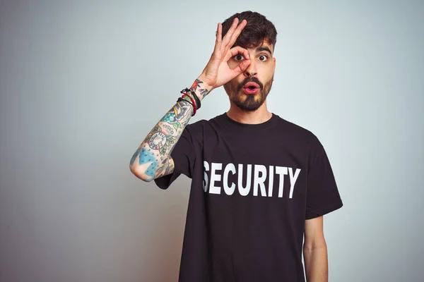 Young safeguard man with tattoo wering security uniform over isolated white background doing ok gesture shocked with surprised face, eye looking through fingers. Unbelieving expression.