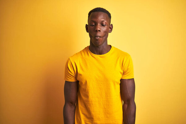 Young african american man wearing casual t-shirt standing over isolated yellow background making fish face with lips, crazy and comical gesture. Funny expression.