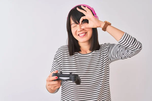 Chinese gamer woman playing video game using headphones over isolated white background with happy face smiling doing ok sign with hand on eye looking through fingers