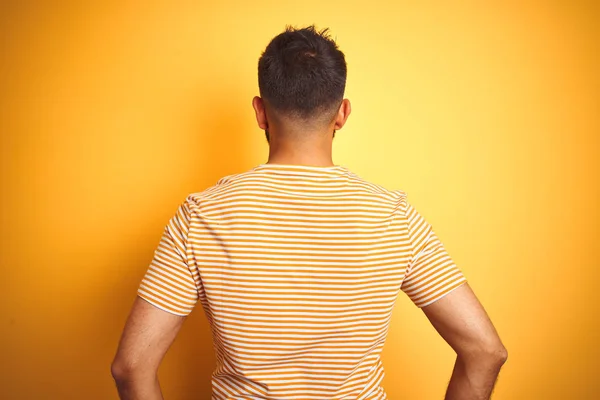 孤立した黄色の背景の上に立つTシャツを着た若いインド人男性が 体に腕を向けて後ろ向きに立っている — ストック写真