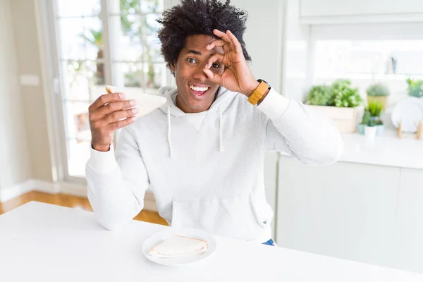 African American man eating handmade sandwich at home with happy face smiling doing ok sign with hand on eye looking through fingers