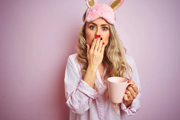 Young woman wearing pajama and mask drinking a cup of coffee over pink isolated background cover mouth with hand shocked with shame for mistake, expression of fear, scared in silence, secret concept