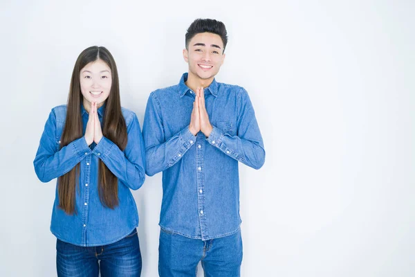 Beautiful young asian couple over white isolated background praying with hands together asking for forgiveness smiling confident.