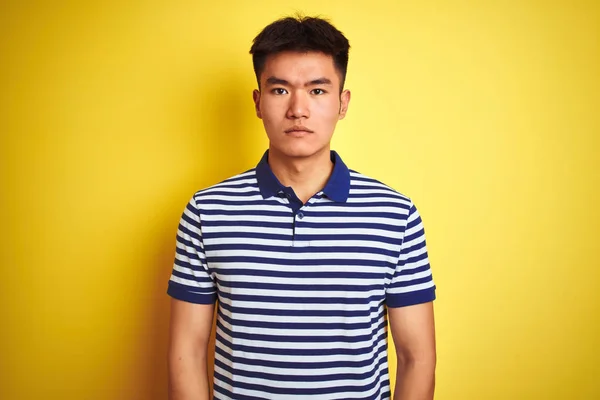 孤立した黄色の背景の上に立つストライプのポロを着た若いアジアの中国人男性は 顔に真剣な表情でリラックスしました カメラを見てシンプルで自然な — ストック写真