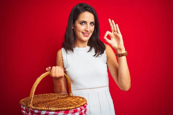 Young beautiful woman on picnic holding wicker basket over red isolated background doing ok sign with fingers, excellent symbol