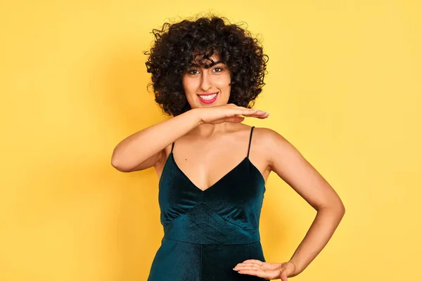 Young arab woman with curly hair wearing elegant dress over isolated yellow background gesturing with hands showing big and large size sign, measure symbol. Smiling looking at the camera. Measuring concept.