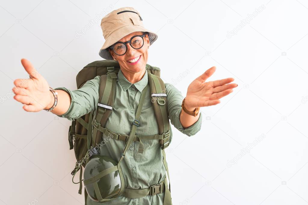 Middle age hiker woman wearing backpack canteen hat glasses over isolated white background looking at the camera smiling with open arms for hug. Cheerful expression embracing happiness.