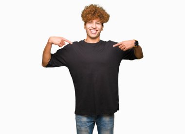 Siyah t-shirt giyen afro saç ile genç yakışıklı adam yüzünde gülümseme ile kendine güvenen görünümlü, parmaklarıyla gurur ve mutlu kendini işaret.