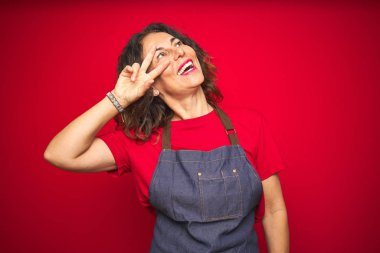 Kırmızı izole arka plan üzerinde önlük üniforma giyen orta yaş yaşlı kadın yüz üzerinde parmakları ile barış sembolü yapıyor, zafer gösteren neşeli gülümseyen