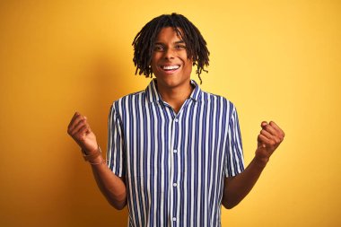 Dreadlocks ile rahat çizgili t-shirt izole sarı arka plan üzerinde sürpriz kutluyor ve kollarını kaldırdı ve açık gözleri ile başarı için şaşırttı giyen Afro adam. Kazanan kavramı.