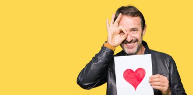 Yakışıklı orta yaş adam mutlu yüz ile kırmızı kalp ile kağıt tutarak gözler parmaklarıyla görünümlü el ile ok işareti yapıyor gülümseyen