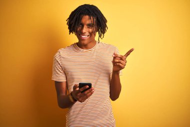 Izole sarı arka plan üzerinde akıllı telefon kullanarak dreadlocks ile Afro Amerikan adam çok mutlu yan el ve parmak ile işaret