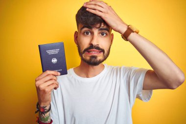 Izole sarı arka plan üzerinde Alman Almanya pasaportu giyen dövme ile genç adam baş el ile vurguladı, utanç ve sürpriz yüz ile şok, kızgın ve sinirli. Korku ve hata için üzgün.