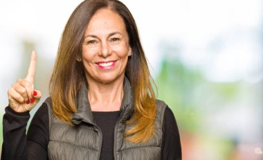 Güzel orta yaş kadın gösteren kış yelek giyen ve parmak bir numara ile işaret kadar emin ve mutlu gülümseyen.