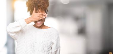 Gülümsüyor ve gözleri için sürpriz kapsayan yüz el ile gülüyor izole arka plan üzerinde kış kazak giyen güzel genç Afro-Amerikan kadın. Kör kavramı.