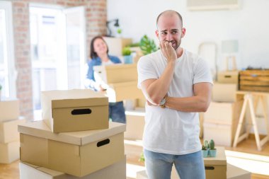 Young couple arround cardboard boxes moving to a new house, bald man standing at home looking stressed and nervous with hands on mouth biting nails. Anxiety problem. clipart