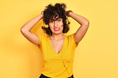 Kıvırcık saçlı Arap kadın izole sarı arka plan üzerinde kulaklık kullanarak müzik dinlerken baş ağrısı umutsuz muzdarip ve stresli çünkü ağrı ve migren. Eller baş üstünde.