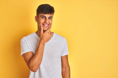 Beyaz tişört giyen genç hintli adam izole sarı arka plan üzerinde duran kamera emin bakarak çapraz kollar ve el çene üzerinde kaldırdı gülümseyerek. Olumlu düşünme.