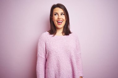 Genç güzel esmer kadın pembe izole arka plan üzerinde bir kazak giyen yan bakarak gülümseyerek ve düşünme uzak bakıyor.