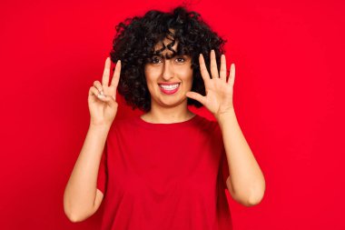 İzole kırmızı arka plan üzerinde rahat t-shirt giyen kıvırcık saçlı genç arap kadın gösteren ve emin ve mutlu gülümserken parmakları ile numara yedi işaret.
