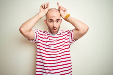 Sakallı genç kel adam beyaz izole arka plan üzerinde rahat çizgili kırmızı t-shirt giyen boğa boynuzları gibi baş üzerinde parmak ile komik jest yapıyor