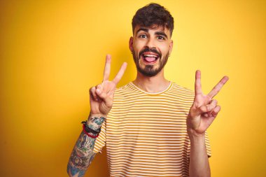 Çizgili t-shirt giyen dövme giyen genç adam izole sarı arka plan üzerinde duran dil ile gülümseyerek her iki elinin parmakları zafer işareti yapıyor gösteren. İki numara.