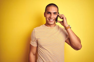 Genç yakışıklı adam ayakta mutlu bir yüz ile sarı izole arka plan üzerinde akıllı telefon kullanarak konuşurken ve dişleri gösteren kendinden emin bir gülümseme ile gülümseyerek