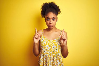 Izole sarı arka plan üzerinde duran rahat çiçekli elbise giyen Afro-amerikan kadın üzgün ve üzgün görünümlü kadar işaret, parmakları ile yön gösteren, mutsuz ve depresif.