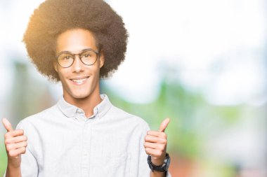 El ile pozitif jest yapıyor gözlük başarı işareti giyiyor afro saçlı genç Afro-Amerikan adam başparmak gülümseyen ve mutlu. Neşeli bir ifadeyle, kazanan jest kameraya bakıyor.