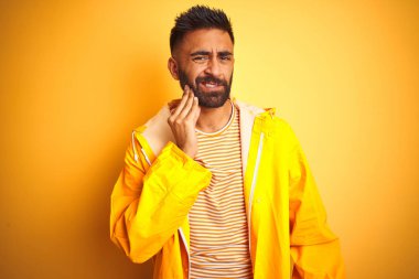 Genç hintli adam izole sarı arka plan üzerinde ayakta yağmurluk giyen diş ağrısı veya diş üzerinde diş hastalığı nedeniyle ağrılı ifade ile el ile ağız dokunmadan. Diş hekimi konsepti.
