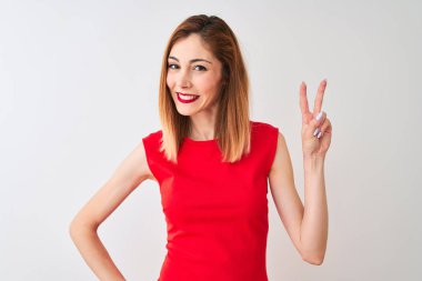 Kırmızı başlı iş kadını izole beyaz arka plan üzerinde duran zarif kırmızı elbise giyen kameraya bakarak gülümseyerek zafer işareti yapıyor parmakları gösteren. İki numara.