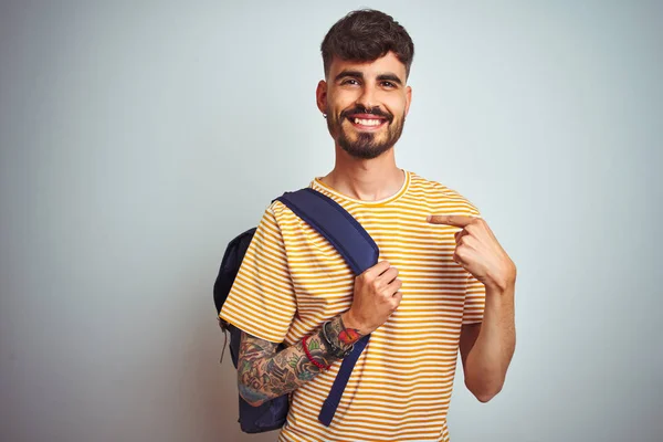 Young student man with tattoo wearing backpack standing over isolated white background with surprise face pointing finger to himself
