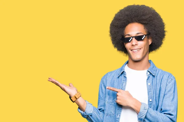 Young african american man with afro hair wearing thug life glasses amazed and smiling to the camera while presenting with hand and pointing with finger.