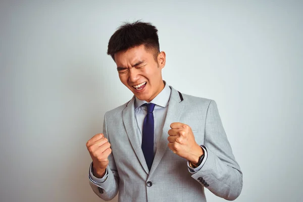 Asian chinese businessman wearing grey jacket and tie standing over isolated white background very happy and excited doing winner gesture with arms raised, smiling and screaming for success. Celebration concept.