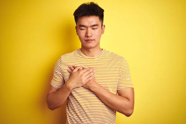 孤立した黄色の背景の上に立つTシャツを着たアジアの若い中国人男性は 目を閉じて感謝のジェスチャーで胸に手を当て 笑顔を浮かべていた 健康の概念 — ストック写真
