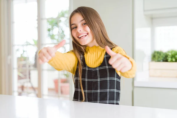 Young beautiful blonde kid girl wearing casual yellow sweater at home approving doing positive gesture with hand, thumbs up smiling and happy for success. Winner gesture.