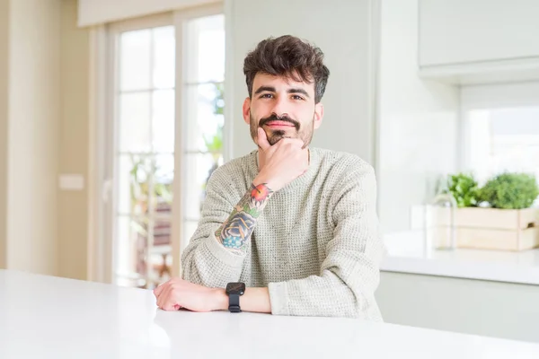 Young man wearing casual sweater sitting on white table looking confident at the camera with smile with crossed arms and hand raised on chin. Thinking positive.