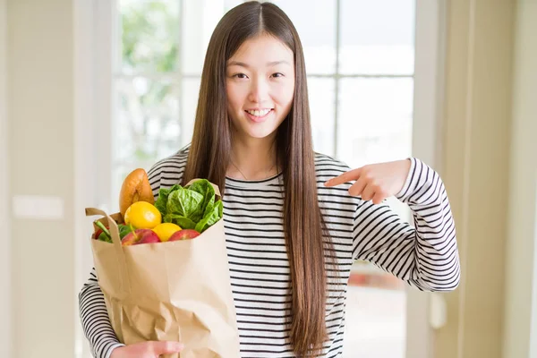 美しいアジアの女性は 自分自身に指を指して驚きの顔で新鮮な食料品の紙袋を保持 — ストック写真