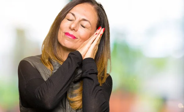 Beautiful middle age woman wearing winter vest sleeping tired dreaming and posing with hands together while smiling with closed eyes.